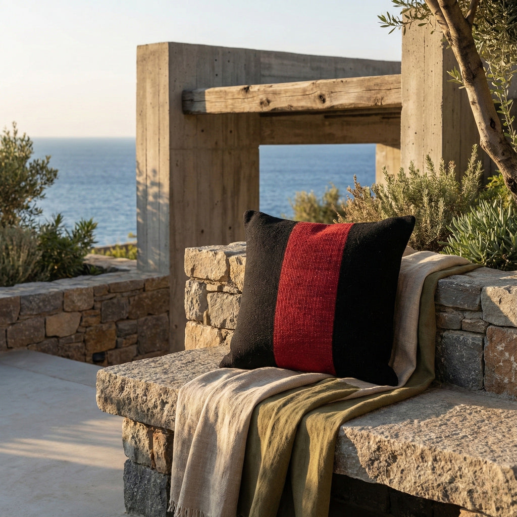 Handwoven black pillow cover with a red stripe on a stone bench, outdoor terrace with sea view.