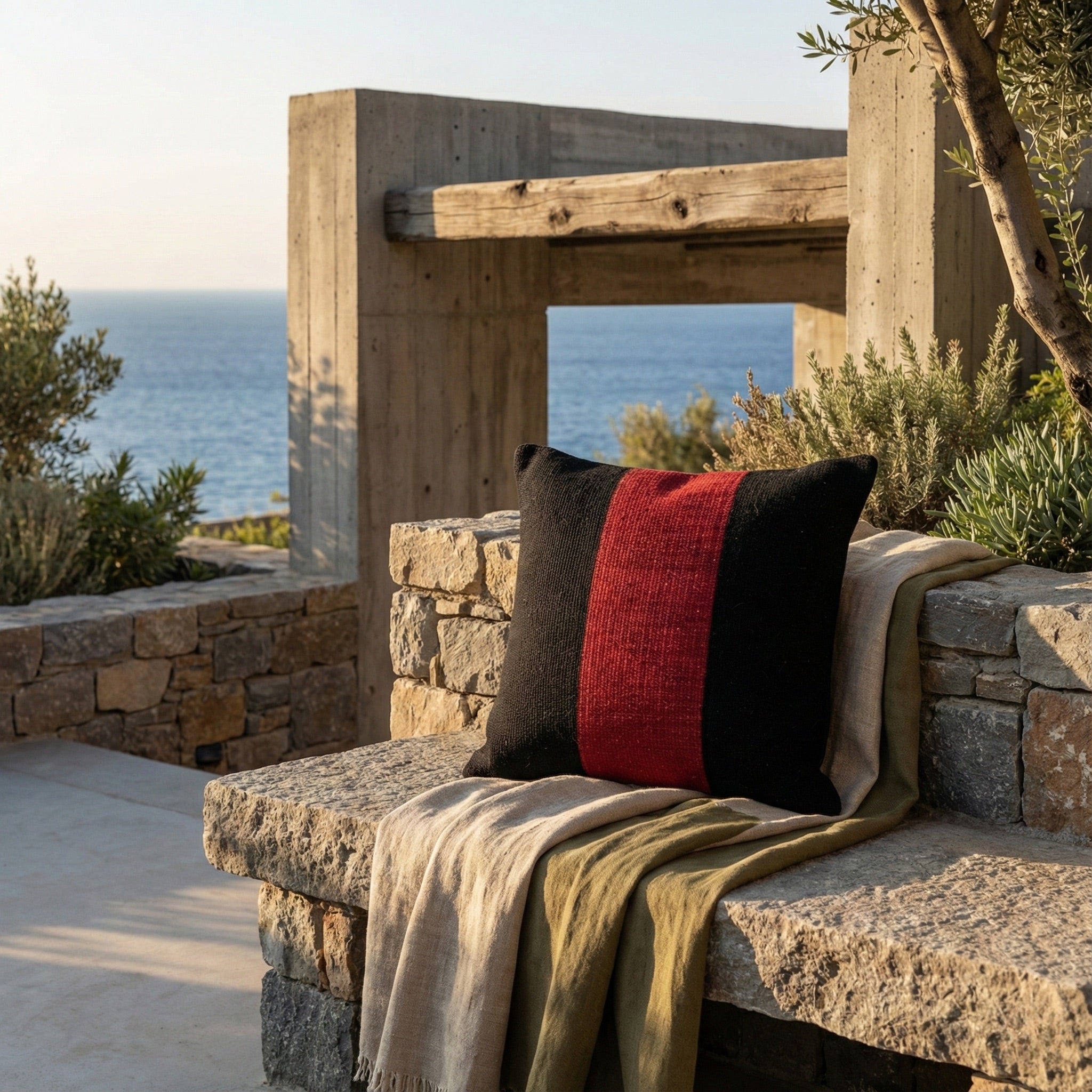 Handwoven black pillow cover with a red stripe on a stone bench, outdoor terrace with sea view.